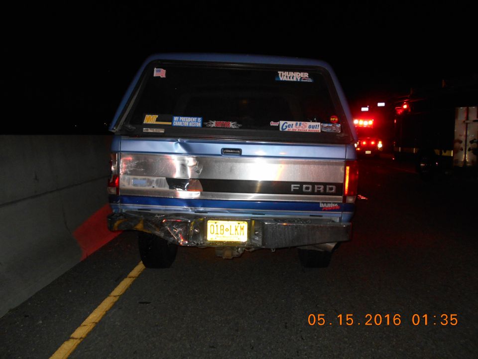Rear of a blue Ford pickup truck with a camper shell, damaged bumper, and stickers on the back window. Another vehicle with flashing lights is visible ahead