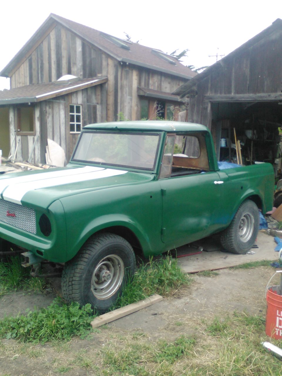 Green International Harvester Scout pickup truck with white racing stripes on the hood, parked outside a wooden building