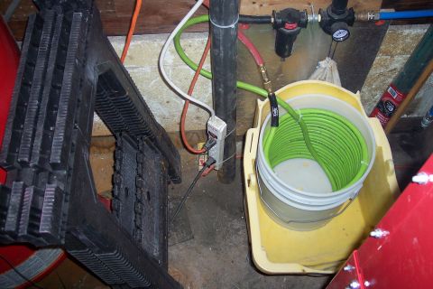 1990 Ford Bronco - An air compressor setup is shown with a coiled green hose in a bucket, a black step stool, and an electrical outlet on a pipe.