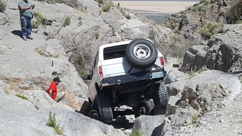 1979 Ford Bronco - A white 1979 Ford Bronco is shown off-roading on a challenging rocky path, with a spare tire mounted on the back.