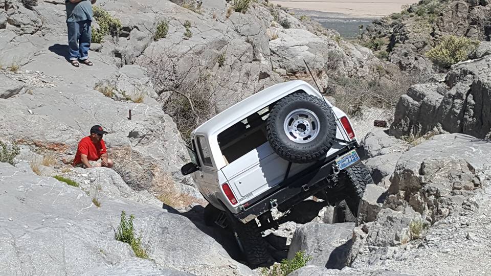 White SUV tilted on a steep, rocky incline with a spare tire mounted on the back. Two people are visible on the rocks nearby