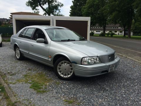 1999 Ford Fairlane - A silver sedan is parked in front of a garage on a gravel lot with trees and buildings in the background.