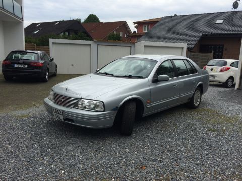 1999 Ford Fairlane - A silver sedan with a distinctive grille and headlights is parked in a driveway next to other vehicles.
