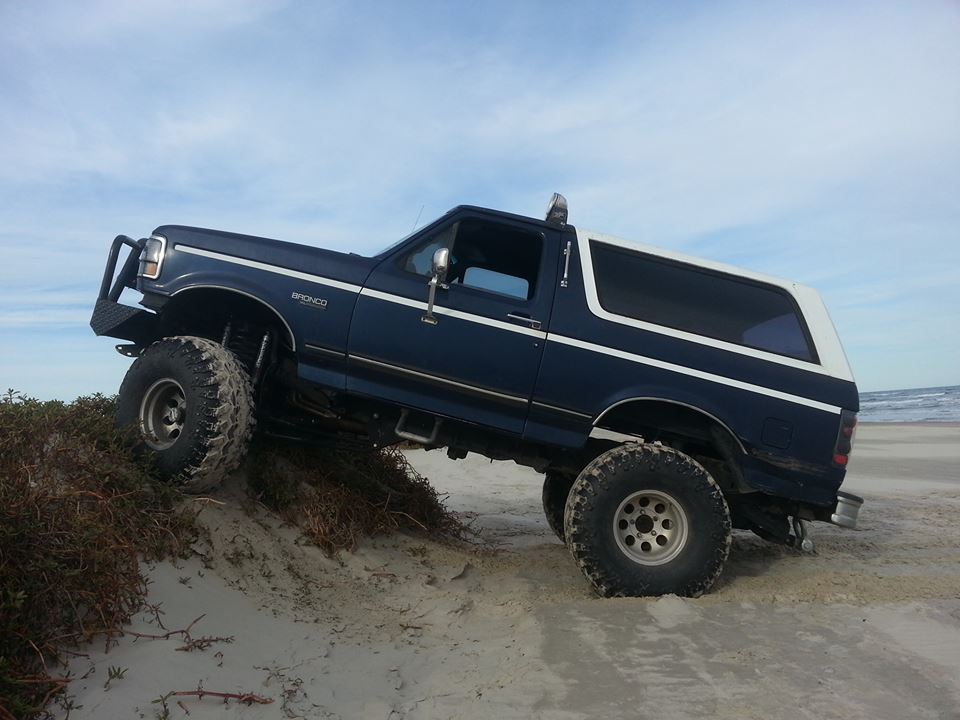 1994 Ford Bronco with large tires climbing a sandy dune near the ocean