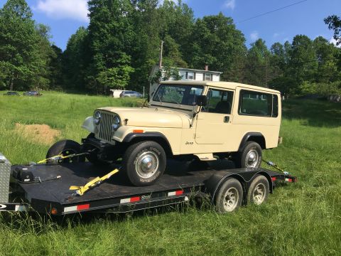 1986 Jeep Wagoneer - A tan Jeep CJ-8 Scrambler is loaded onto a trailer in a grassy field.