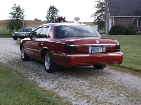 1999 Mercury Grand Marquis - A red sedan is parked on a gravel driveway next to a house, with another car in the background.