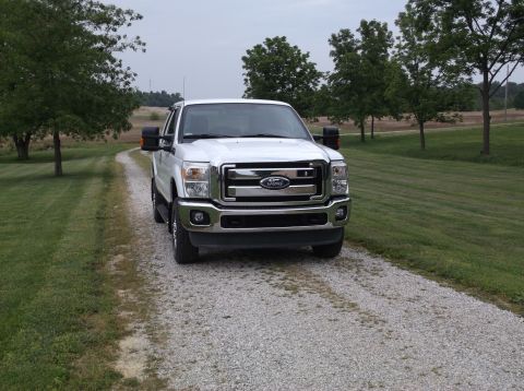2011 Ford F-250 - A white 2011 Ford F-250 is parked on a gravel driveway surrounded by grass and trees.
