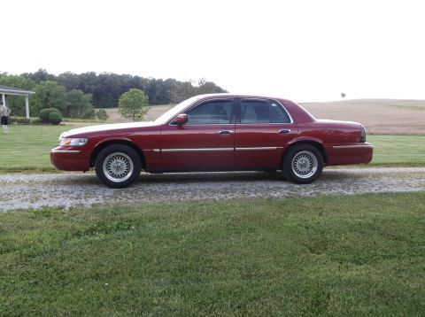 1999 Mercury Grand Marquis - A red sedan is parked on a gravel driveway next to a grassy area with a house and trees in the distance.