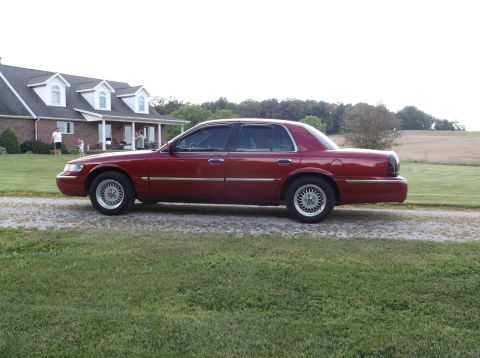 1999 Mercury Grand Marquis - A red sedan is parked on a gravel driveway outside a house.