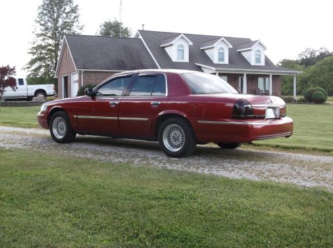 1999 Mercury Grand Marquis - A red 1999 Mercury Grand Marquis is parked on a gravel driveway in front of a house, with a white pickup truck visible to the left.