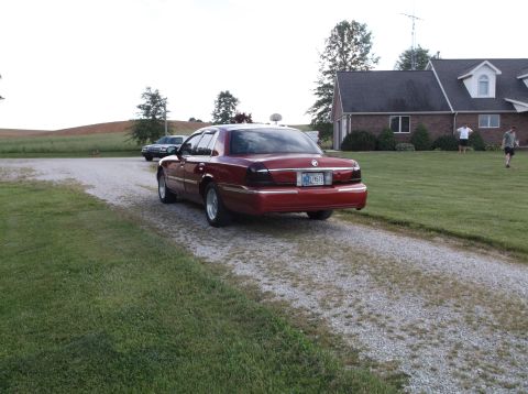 1999 Mercury Grand Marquis - A red sedan is parked on a gravel driveway near a house.
