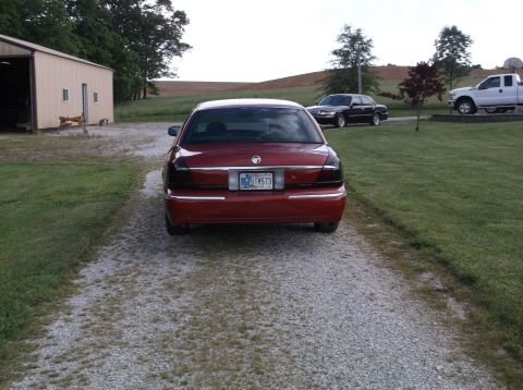 1999 Mercury Grand Marquis - Rear view of a red Mercury Grand Marquis on a gravel driveway, with other vehicles and a building in the background.