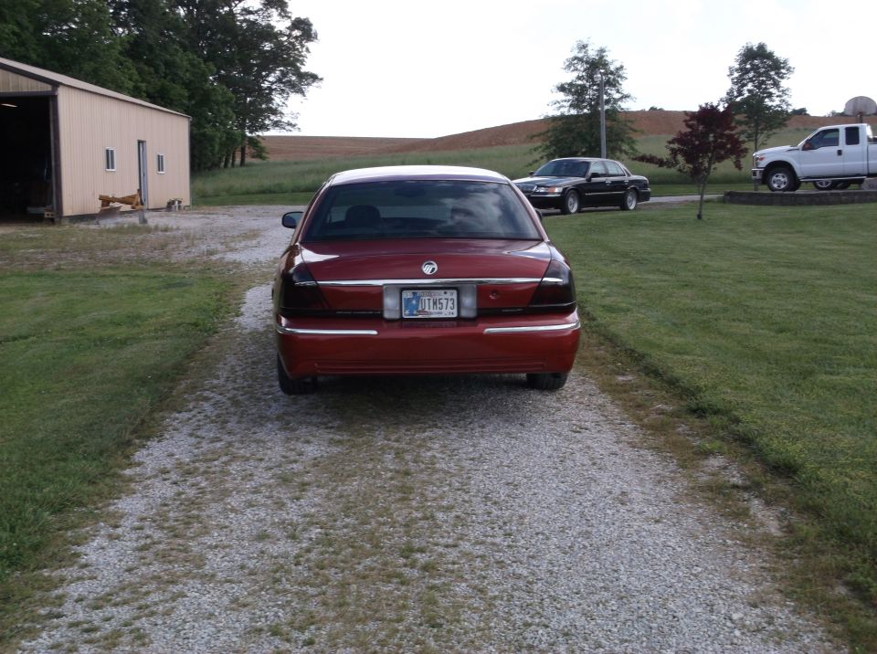 Red Mercury Grand Marquis parked on gravel driveway, rear view. Tinted taillights, chrome trim. Other vehicles and a building visible