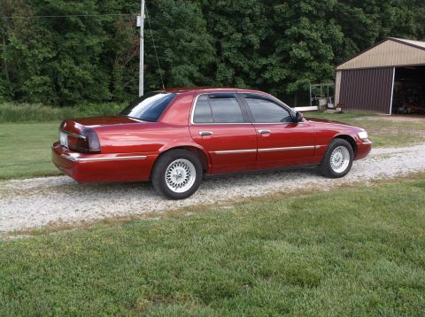 1999 Mercury Grand Marquis - Side view of a red 1999 Mercury Grand Marquis parked on a gravel driveway next to a grassy area.