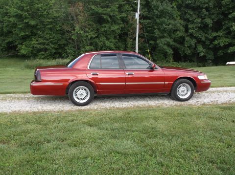 1999 Mercury Grand Marquis - Side view of a red 1999 Mercury Grand Marquis parked on a gravel driveway with grass and trees in the background.