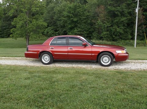 1999 Mercury Grand Marquis - A red 1999 Mercury Grand Marquis sedan is seen from the side, parked on a gravel driveway with grass and trees in the background.
