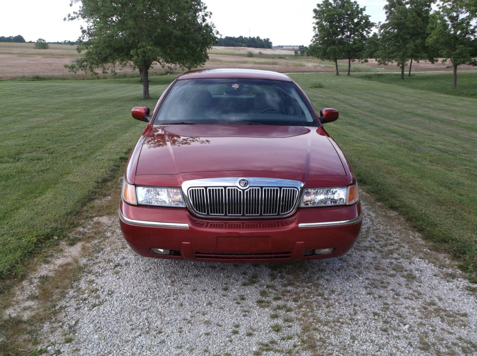 Red Mercury Grand Marquis parked on gravel, front view. Chrome grille and headlights visible