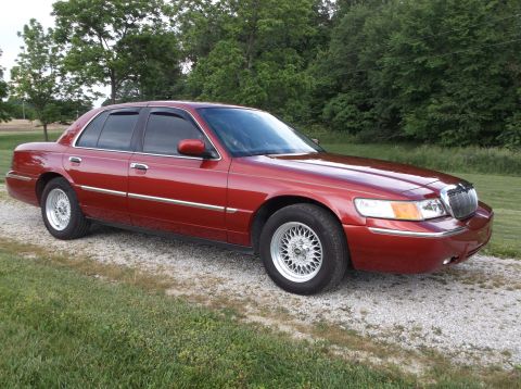 1999 Mercury Grand Marquis - This red 1999 Mercury Grand Marquis is shown from a three-quarter front angle, parked on a gravel surface with trees in the background.