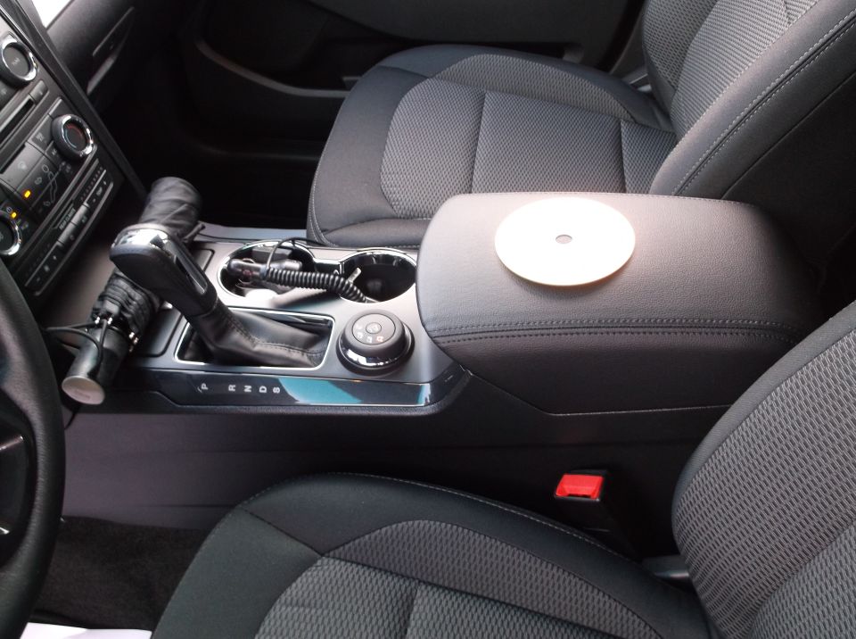 Interior shot of a car's center console with gear shifter, cup holders, and armrest. A white disc sits on the armrest