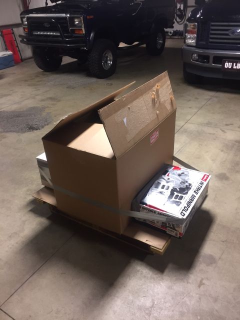 1982 Ford Bronco - A box labeled "INTAKE MANIFOLD" sits on a pallet in front of two trucks.