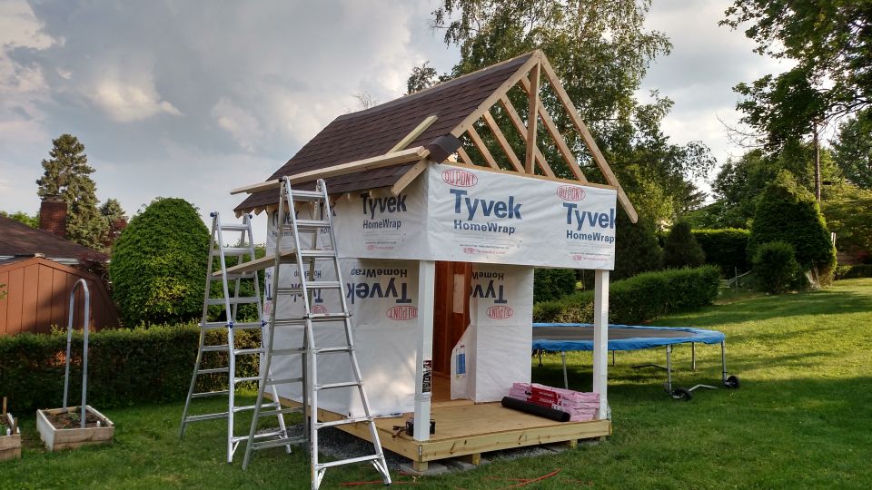 Playhouse under construction with Tyvek wrap, ladders, and roofing materials visible