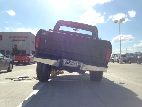 1979 Ford Bronco - This lifted Ford pickup truck is parked outside a Dodge dealership, showing its rear end and custom license plate.