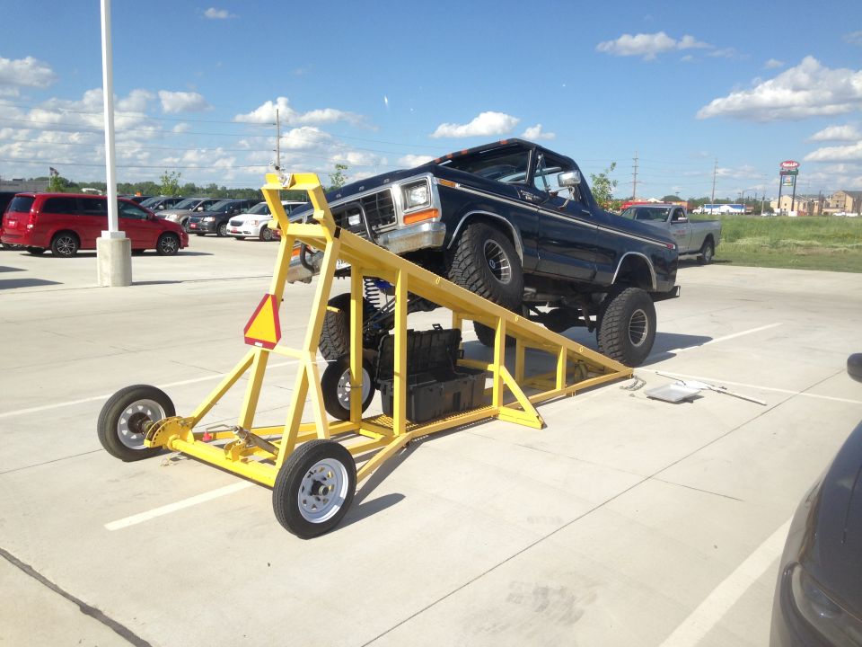 Black 1978 Ford Bronco with lifted suspension on a yellow wheeled ramp. Other vehicles and buildings in background