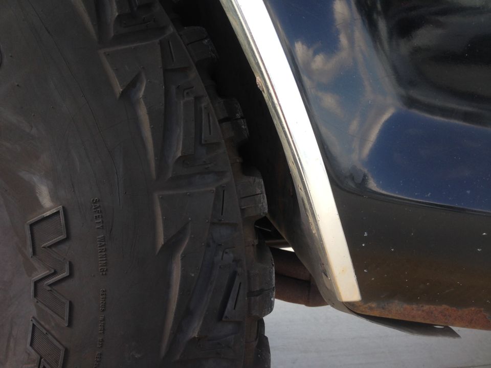 Close-up of a large tire and the lower fender area of a dark-colored vehicle. Some rust is visible on the undercarriage