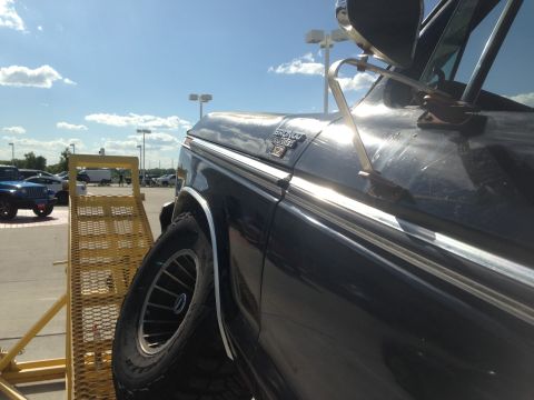 1979 Ford Bronco - A dark-colored Ford Bronco is partially visible, positioned next to a yellow ramp in a parking lot under a partly cloudy sky.