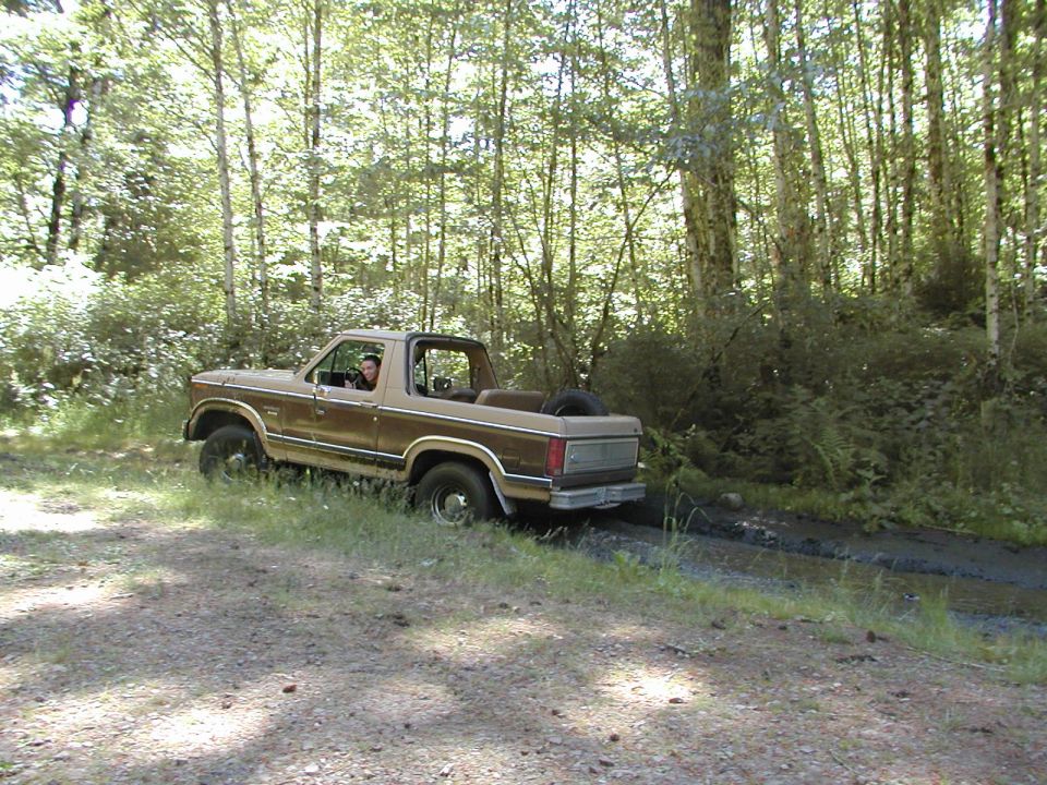 Brown Ford Bronco driving on a dirt path through a forest, with a spare tire mounted on the back