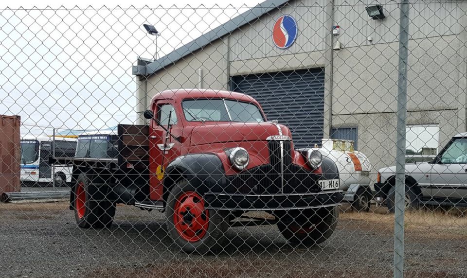 Red vintage truck with black fenders and red wheels parked in front of a building. License plate reads 91 M16