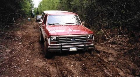 1985 Ford Bronco - A red Ford Bronco navigates a muddy, wooded trail with another vehicle following behind.
