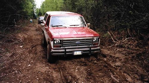 Red Ford pickup with brush guard stuck in a muddy wooded trail, front view