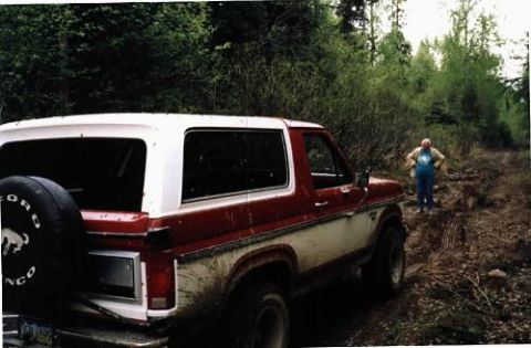 1985 Ford Bronco - A red and white 1985 Ford Bronco is parked on a muddy dirt road surrounded by trees, with a person standing in the background.