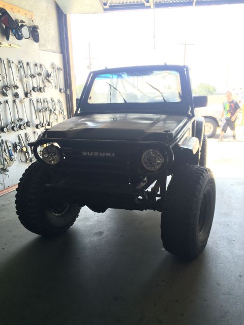 1987 Suzuki Samurai - Front view of a black 1987 Suzuki Samurai modified with oversized tires and auxiliary lighting, parked indoors.