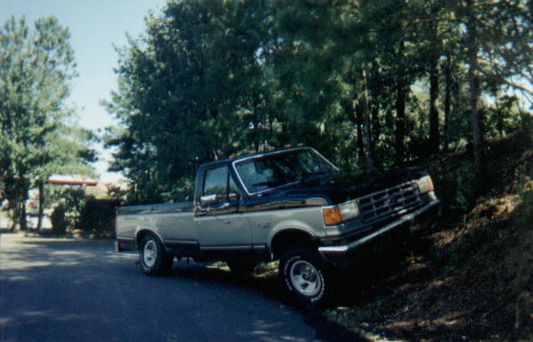 Two-tone dark and silver Ford regular-cab pickup parked beside a tree-lined driveway, front 3/4 view