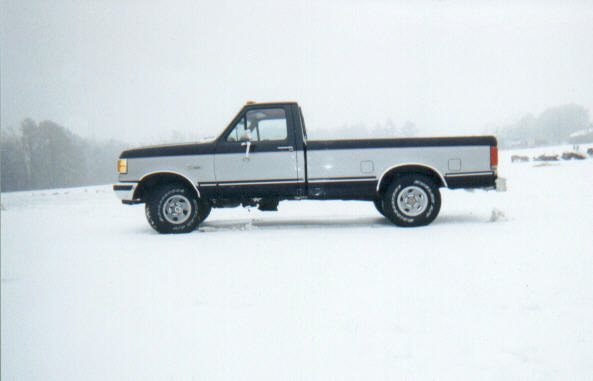 Side view of two-tone blue and silver Ford pickup parked in a snowy, foggy field
