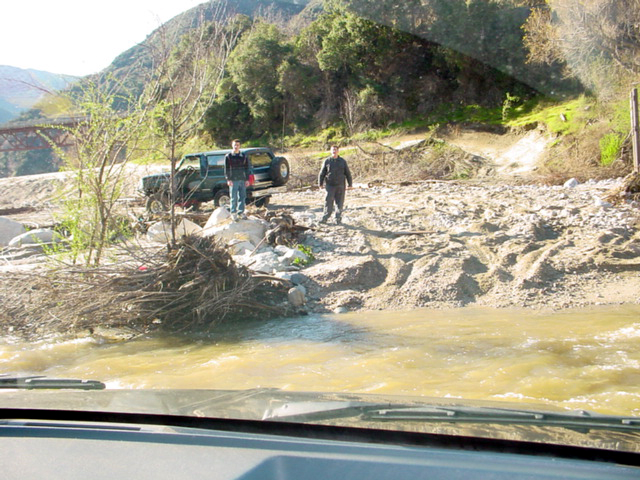 Two men stand by a green SUV parked on a rocky riverbed with a bridge in the background