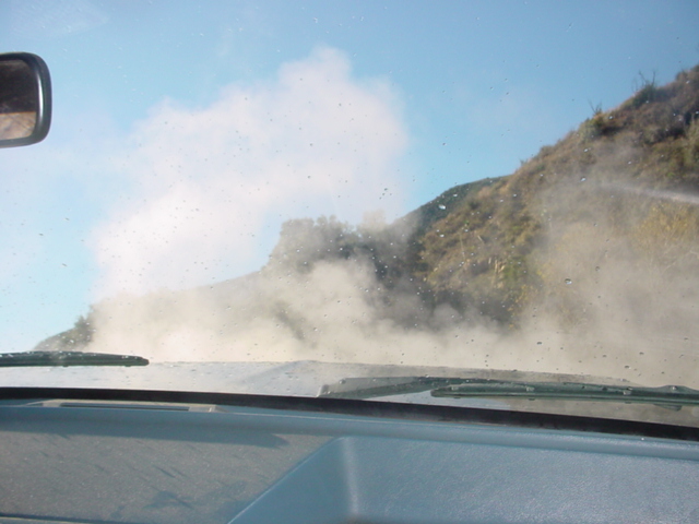 View through a dusty windshield of a dirt road with a large dust cloud rising beside a hill