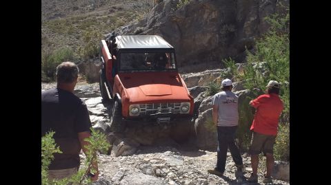 1979 Ford Bronco - An orange off-road vehicle with a white soft top is shown on a rocky trail with three onlookers.