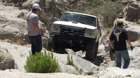 1979 Ford Bronco - A white Ford Bronco is shown off-roading through a rocky landscape with two onlookers present.