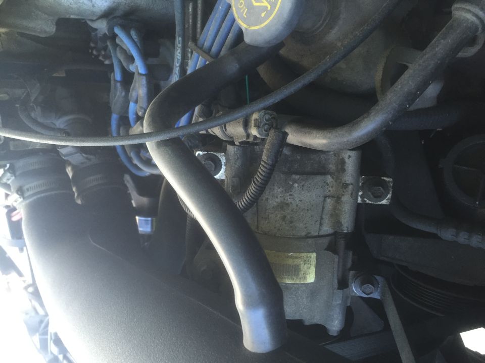 Close-up of an engine component, possibly an AC compressor, with hoses and wiring