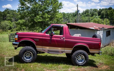 1995 Ford Bronco - A lifted 1995 Ford Bronco in a rural setting, showing its off-road modifications and a weathered outbuilding.