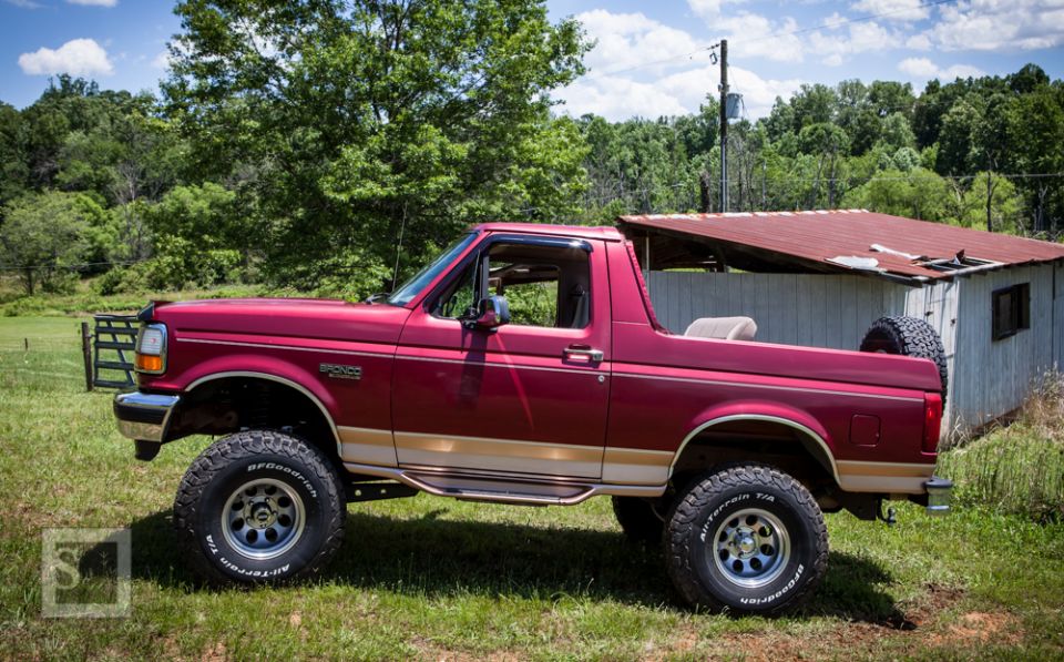 Red 1995 Ford Bronco with tan trim, lifted on off-road tires, parked in a grassy field next to a dilapidated shed