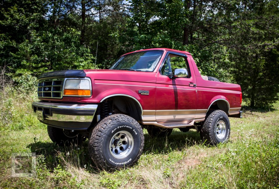 1995 Ford Bronco, red, lifted with BFGoodrich All-Terrain T/A tires, parked in grassy area with trees in background