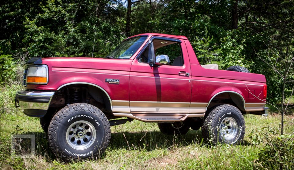 Red Ford Bronco with tan trim and off-road tires parked in grassy area with trees