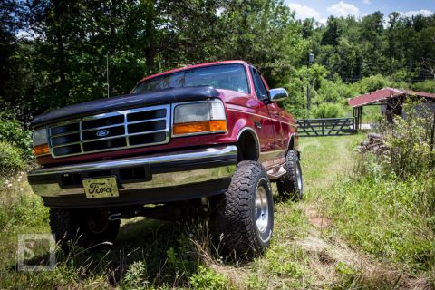 1995 Ford Bronco - A lifted red Ford pickup truck sits on a grassy path near a wooden fence and a small building.