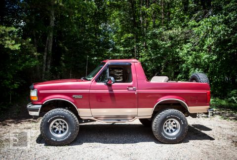1995 Ford Bronco - This red Ford Bronco is parked outdoors with trees in the background. It has a spare tire in the cargo area.