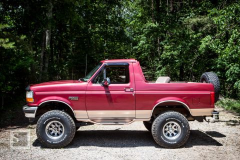 1995 Ford Bronco - A red 1995 Ford Bronco is parked outdoors, showing its side profile with off-road tires and a spare tire on the back.