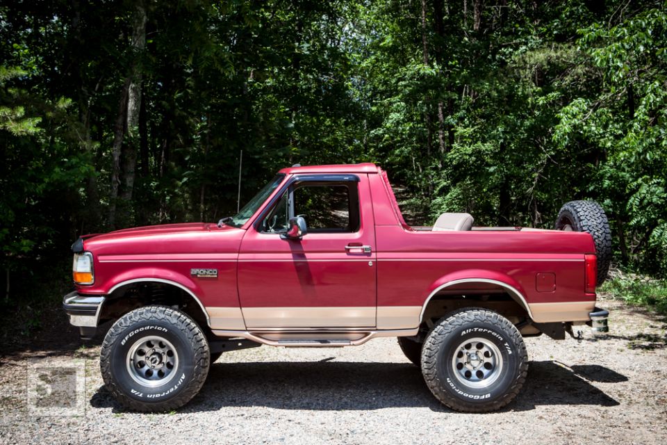 Red 1995 Ford Bronco with tan lower body trim and a spare tire mounted on the rear, viewed from the side